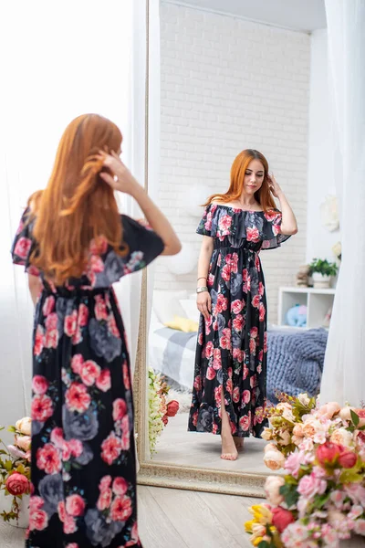 Attractive young woman in floral print dress stands in front large mirror in graceful floral interior. pretty girl with curly red hair, in beautiful dress, in floral interior, smiles. spring festival