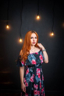 Güzel, çarpıcı, kızıl saçlı bir kadın çiçek desenli siyah bir elbisenin içinde dikiliyor. Siyah arka planda dikiliyor, ayna ve parlayan ampullerle. Gece makyajlı çekici kız ileriye bakıyor.