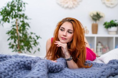 Kızıl saçlı çekici bir kadının portresi. Yatağın üzerinde büyük örülmüş merino yününden yapılmış gri bir battaniyenin üzerinde yatıyor. Cilt bakımı, şehvet ve kadınsı güzellik kavramı..