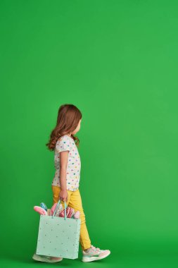 Paskalya tavşanı kulakları takan şık elbiseli şirin küçük kız, Paskalya gününde yeşil arka planda tavşan kulaklı bir çantayla gider. Tam boy. Boşluğu kopyala.