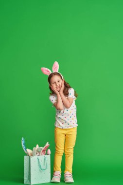 Paskalya tavşanı kulakları takan şık elbiseli şirin küçük kız, Paskalya gününde yeşil arka planda tavşan kulaklı bir çantayla gider. Tam boy. Boşluğu kopyala.