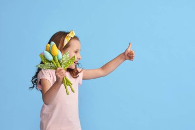 Saçları kıvırcık ve kafasında bandaj olan küçük tatlı kız renkli laleler tutuyor. Çocuk gülümsüyor ve mavi arka plan stüdyosunda işaret onayını gösteriyor. konsept Paskalya ve Bahar
