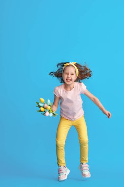 Kıvırcık saçlı, buket renkli laleli güzel bir kız mavi arka plan stüdyosunda boyuna kadar zıpladı. Moda kıyafetleri içinde bir çocuk. Dinamik resim