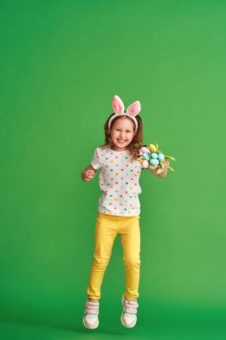 Paskalya tavşanı kulaklı sevimli mutlu kız yeşil bir arka plana atlıyor. Paskalya Günü 'nde bir çocuk gülümser ve elinde bir buket kumaş lale çiçeği tutar. Baharın gelişinin neşesi.