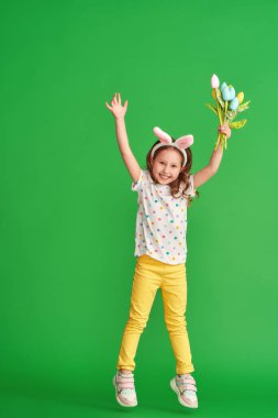 Paskalya tavşanı kulaklı sevimli mutlu kız yeşil bir arka plana atlıyor. Paskalya Günü 'nde bir çocuk gülümser ve elinde bir buket kumaş lale çiçeği tutar. Baharın gelişinin neşesi.