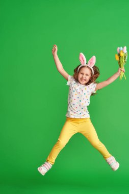 Paskalya tavşanı kulaklı sevimli mutlu kız yeşil bir arka plana atlıyor. Paskalya Günü 'nde bir çocuk gülümser ve elinde bir buket kumaş lale çiçeği tutar. Baharın gelişinin neşesi.