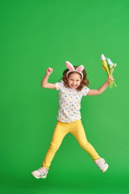 Paskalya tavşanı kulaklı sevimli mutlu kız yeşil bir arka plana atlıyor. Paskalya Günü 'nde bir çocuk gülümser ve elinde bir buket kumaş lale çiçeği tutar. Baharın gelişinin neşesi.