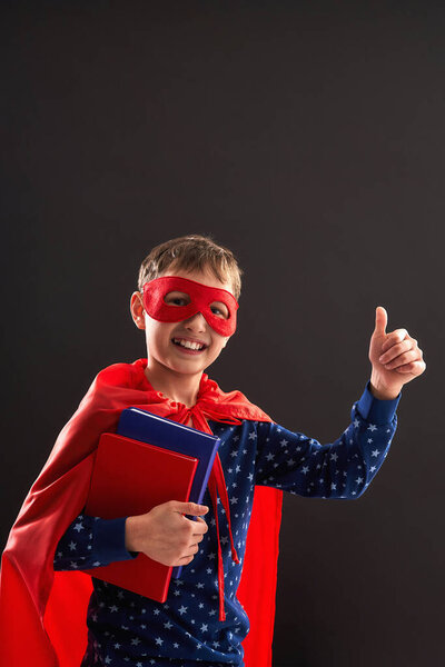 cheerful child, dressed in superhero costume, holds books in his hand and shows class, sign approval. little boy laughs and enjoys going to school. Posing on a black background in studio.