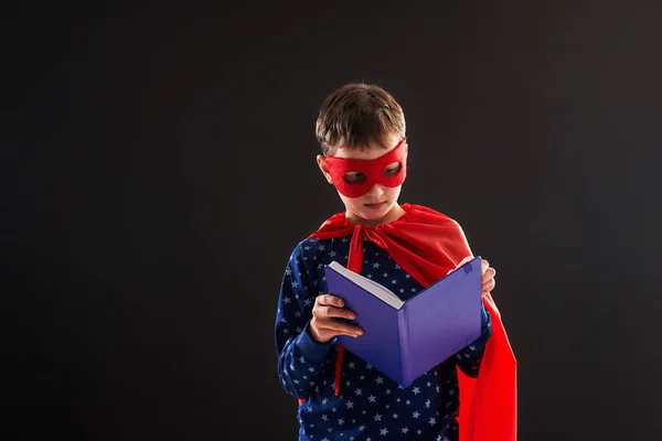 attentive child, dressed in superhero costume, holds books in hand and reads enthusiastically. boy is interested in new book, he is happy to get knowledge. Learn to read quickly on black background.