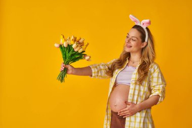 Çekici, hamile bir sarışın Paskalya günü tavşan kulağı takar ve sarı arka planda duran buket laleleri tutar. Hamile anne elini karnına koyar. Baharda bebek bekliyor..