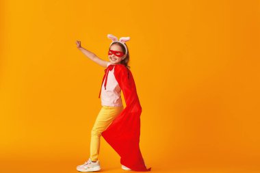 Eğlenceli küçük kız süper kahraman kostümüyle oynuyor. Bebek tavşan kulağı takmış, kırmızı pelerin, maske takmış sarı arka planda elini uzatıyor. konsept kızın süper güçleri, feminizm, zafer için çabalama