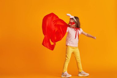 Eğlenceli küçük kız süper kahraman kostümüyle oynuyor. Bebek tavşan kulağı takmış, kırmızı pelerin, maske takmış sarı arka planda elini uzatıyor. konsept kızın süper güçleri, feminizm, zafer için çabalama