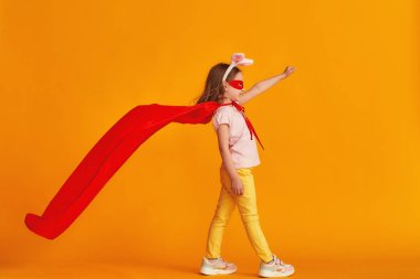 Eğlenceli küçük kız süper kahraman kostümüyle oynuyor. Bebek tavşan kulağı takmış, kırmızı pelerin, maske takmış sarı arka planda elini uzatıyor. konsept kızın süper güçleri, feminizm, zafer için çabalama