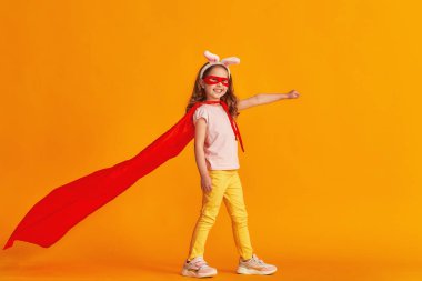 Eğlenceli küçük kız süper kahraman kostümüyle oynuyor. Bebek tavşan kulağı takmış, kırmızı pelerin, maske takmış sarı arka planda elini uzatıyor. konsept kızın süper güçleri, feminizm, zafer için çabalama