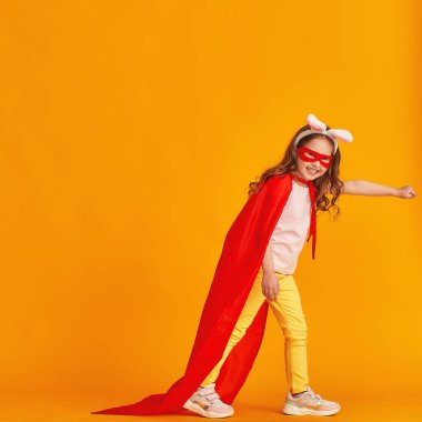 Eğlenceli küçük kız süper kahraman kostümüyle oynuyor. Bebek tavşan kulağı takmış, kırmızı pelerin, maske takmış sarı arka planda elini uzatıyor. konsept kızın süper güçleri, feminizm, zafer için çabalama