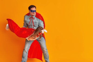 Sakallı ciddi bir baba, küçük bir kızı elinde tutan süper kahraman, Süpermen kostümleri içinde, sarı bir duvarda izole edilmiş bir çocuk partisine hazırlanıyor. Güçlü kahramanlar