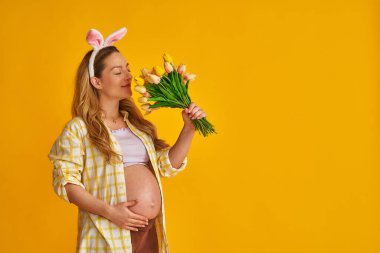 Çekici, hamile bir sarışın Paskalya günü tavşan kulağı takar ve sarı arka planda duran buket laleleri tutar. Hamile anne elini karnına koyar. Baharda bebek bekliyor..