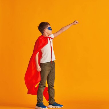 Neşeli çocuk süper kahraman kostümüyle oynuyor. Çocuk kırmızı bir pelerin ve maske takmış, sarı bir arka planda elini uzatıyor. Zafer için çabalayan süper güçlü çocuk konsepti