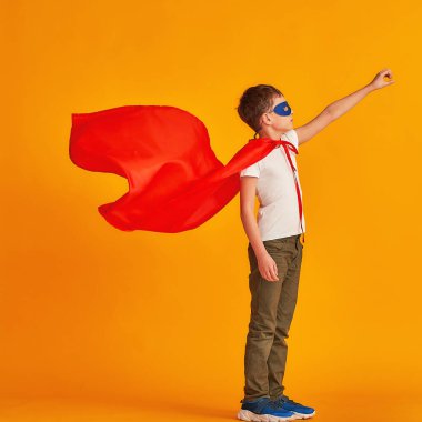 Neşeli çocuk süper kahraman kostümüyle oynuyor. Çocuk kırmızı bir pelerin ve maske takmış, sarı bir arka planda elini uzatıyor. Zafer için çabalayan süper güçlü çocuk konsepti