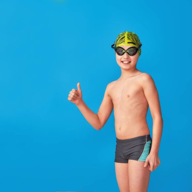 Mavi arka planda mayo, gözlük ve mayo takan küçük bir yüzücü. Çocuk onaylıyor, onaylıyor, reklam yapıyor. Oğlan bir spor yüzme okulunda okuyor. Boşluğu kopyala
