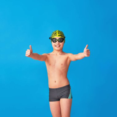Mavi arka planda mayo, gözlük ve mayo takan küçük bir yüzücü. Çocuk onaylıyor, onaylıyor, reklam yapıyor. Oğlan bir spor yüzme okulunda okuyor. Boşluğu kopyala