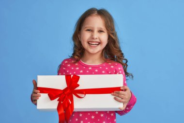 Mavi arka planda kırmızı kurdeleli hediye kutusu olan küçük şirin bir kız. Doğum günü çocuğu. Mutlu çocuk hediyeyi elinde tutuyor. Doğum günü hediyesi. Kutlama, verme ve alma konsepti