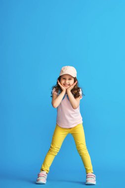 Şık yaz kıyafetleri ve kulaklıkları içinde küçük şirin bir kız mavi bir arka planda poz veriyor. Yaz ve bahar çocuk kıyafetleri konsepti.