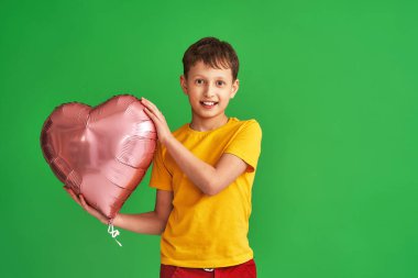 8 yaşında mutlu, gülümseyen bir çocuk, sarı tişört giyiyor, kalbinde pembe folyo topuyla yeşil arka planda duruyor. İlk aşk, aşık olmak ve Sevgililer Günü konsepti.