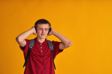 Sırt çantalı ve kulaklıklı mutlu genç bir öğrenci sarı arka planda gözleri kapalı müzik dinliyor. İnsanların yaşam tarzı kavramı. Kopya alanı..