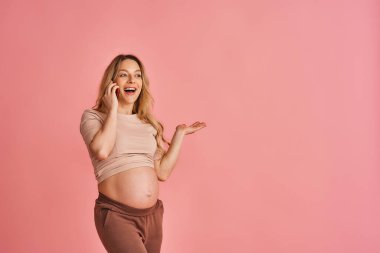 Mutlu hamile kadın, yeni modern akıllı telefondan konuşuyor ve iletişim kuruyor ve elini pembe arka planda yan yana gösteriyor. Müstakbel anne arkadaşlarıyla iletişim kurar ve iyi telefon bağlantılarından hoşlanır.