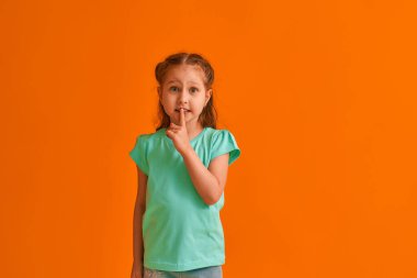 Sersemlemiş, korkmuş, işaret parmağını ağzına dayayan küçük bir kızın portresi turuncu bir arka planda duruyor. - Çocuk konuşmak istemiyormuş. Sır saklar, sır öğrenir.