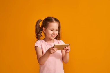 İki at kuyruklu sevimli bir kız stüdyoda turuncu bir arka planda modern bir akıllı telefon tutuyor. İnternete ve cihazlara bağımlı. Yeni bir akıllı telefon reklamı..