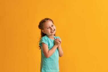 Sevimli küçük kız ellerini göğsünde tutuyor, minnettar hissediyor, çocuk dost canlısı bir ifadeyle avuçlarını göğsüne bastırıyor. Sarı-turuncu arka planda aşk ve inanç kavramı