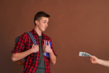 Sırt çantasıyla kitap tutan mutlu genç bir öğrenci kahverengi arka planda ona para sayarken el ele bakıyor. Dolandırıcılık, ek kazanç. konsept üniversite