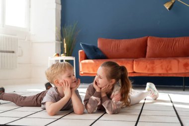 Küçük mutlu beyaz çocuklar, 5 yaşında bir oğlan ve kız, rahat kıyafetler giyip, halıya uzanıp birbirleriyle iletişim kuruyorlar. Mutlu bir çocukluk. Reklam.