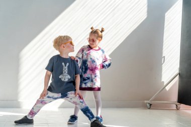 Küçük çekici çocuklar, güneş gözlüklü bir oğlan ve bir kız, şık kıyafetler giyiyorlar, güneşte gülümseyen beyaz bir duvara karşı duruyorlar. Moda reklamları.