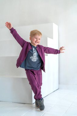 Moda kıyafetleri içinde, gülümseyen, poz veren, koşan ve stüdyonun arka planındaki beyaz duvara atlayan küçük mutlu çocuk. Çocuk kıyafetleri reklamcılığı, duygusal çocuklar, dinamik imaj