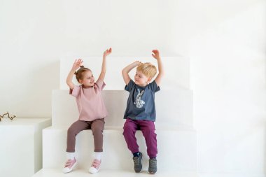 Şık giyinmiş küçük mutlu çocuk ve kız gülümsüyor, beyaz duvar stüdyosuna karşı poz veriyor, merdivenlerde oturuyor. Çocuk kıyafetlerinin reklamını yapmak, duygusal çocuklar, dinamizm imajı. Boşluğu kopyala