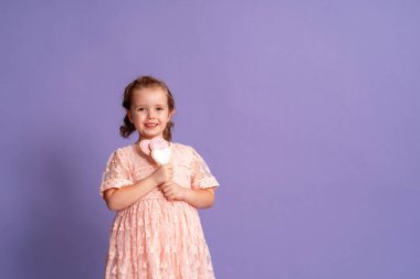 Yumuşak pembe elbiseli gülümseyen küçük kız stüdyoda mor arka planda kalp şeklinde bir çubukta zencefilli ekmek tutuyor. Çocukların doğum günü ve Sevgililer Günü için ikramlar konsepti