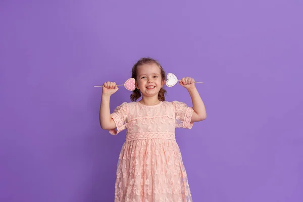 穿着柔软粉色衣服的微笑的小女孩把姜饼放在手杖上 在工作室的紫色背景上 呈心形 儿童节和情人节礼物的概念 — 图库照片