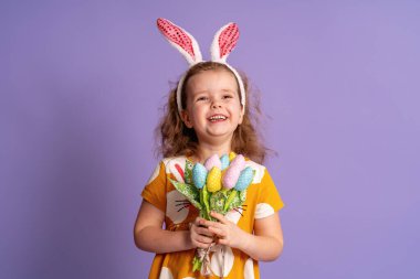 Paskalya tavşanı kulakları ve çiçek desenli bahar lale çiçekleri olan sevimli mutlu küçük bir kız. Mor arka planda bel hizasına kadar kumaş yapmış. Çocuk bahar ve Paskalya geldiğinde gülümser ve sevinir.