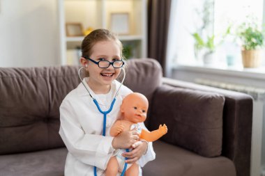 Gözlüklü beyaz bir kız ve bir bebeği tedavi etmek için beyaz tıbbi üniformalı bebek doktorculuk oynuyor. Mutlu bir çocuk, çocuk kliniğinde oyuncağı inceleyen bir doktor gibi davranır..
