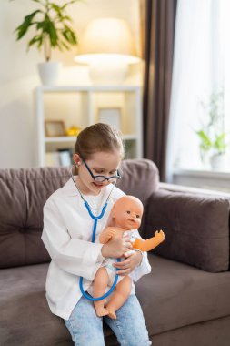 Gözlüklü beyaz bir kız ve bir bebeği tedavi etmek için beyaz tıbbi üniformalı bebek doktorculuk oynuyor. Mutlu bir çocuk, çocuk kliniğinde oyuncağı inceleyen bir doktor gibi davranır..