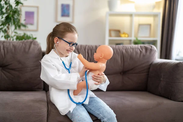 little Caucasian girl in glasses and a white medical uniform to treat a baby doll is playing doctor and hospital. A happy child acts as a doctor examining a toy in a children's clinic.