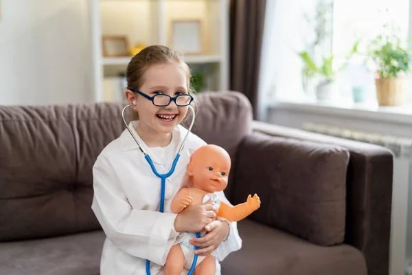 little Caucasian girl in glasses and a white medical uniform to treat a baby doll is playing doctor and hospital. A happy child acts as a doctor examining a toy in a children\'s clinic.