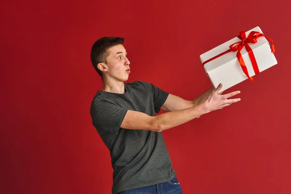 令人惊讶的年轻人 一个身穿休闲装的震惊的白人青少年抓住了一个大的白色礼品盒 上面有一条红色的带子 背景是红色的 赢钱的概念 — 图库照片