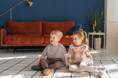 Küçük mutlu beyaz çocuklar, 5 yaşında erkek ve kız, rahat kıyafetler giyiyorlar, halıya oturup birbirleriyle iletişim kuruyorlar. Mutlu bir çocukluk. Reklam.