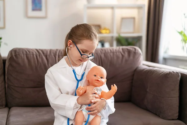 little Caucasian girl in glasses and a white medical uniform to treat a baby doll is playing doctor and hospital. A happy child acts as a doctor examining a toy in a children's clinic.