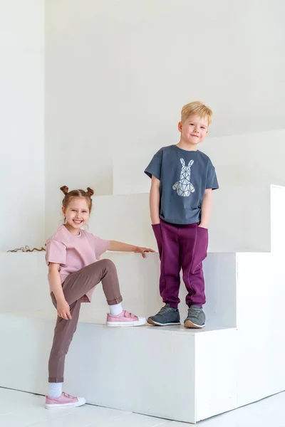 快乐的小男孩和小女孩穿着时髦的衣服 面带微笑 在背景的白墙工作室里摆姿势 儿童服装广告的概念 情感儿童 动态形象 复制空间 — 图库照片