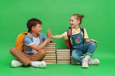 İki mutlu okul çocuğu, bol elbiseli okul kitaplarının üzerine oturur, kitap okur, neşeli bir şekilde gülümser ve yeşil bir arka planda birbirlerine beşlik çakarlar. Ebeveynlik kavramı. Okula geri dön.